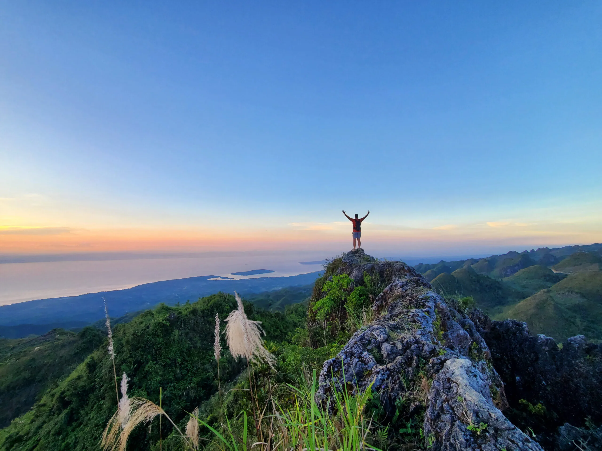 Tim standing on a ledge with his arms raised towards the sunset ocean view from the top of Casino Peak in Cebu, Philippines.