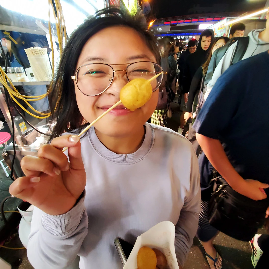Zoe smiling and eating a fried fish ball on a wooden skewer at the Zhongli Night Market in Taipei, Taiwan.