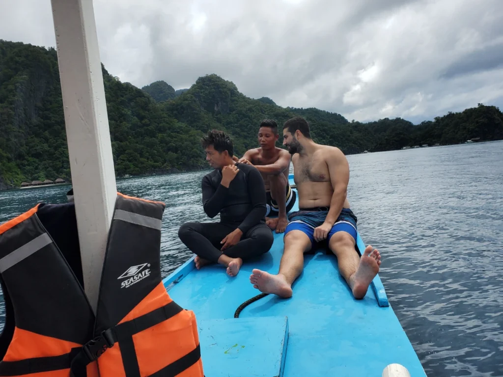Tim sitting on the bow of a blue boat conversing with Filipino locals as they show him a battle wound scar from a box jellyfish. Making meaningful connections while traveling is a favorite among the many benefits of slow travel.