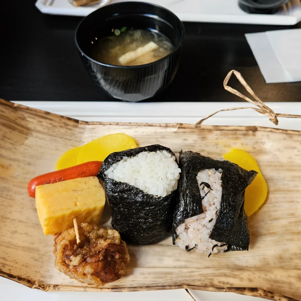 Breakfast of onigiri, tamagoyaki, hotdog, karaage, and pickled radish.