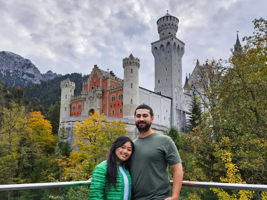 Tim and Zoe standing near the magnificent Neuschwanstein Castle. The castle is orange and white, surrounded by yellow and green trees.