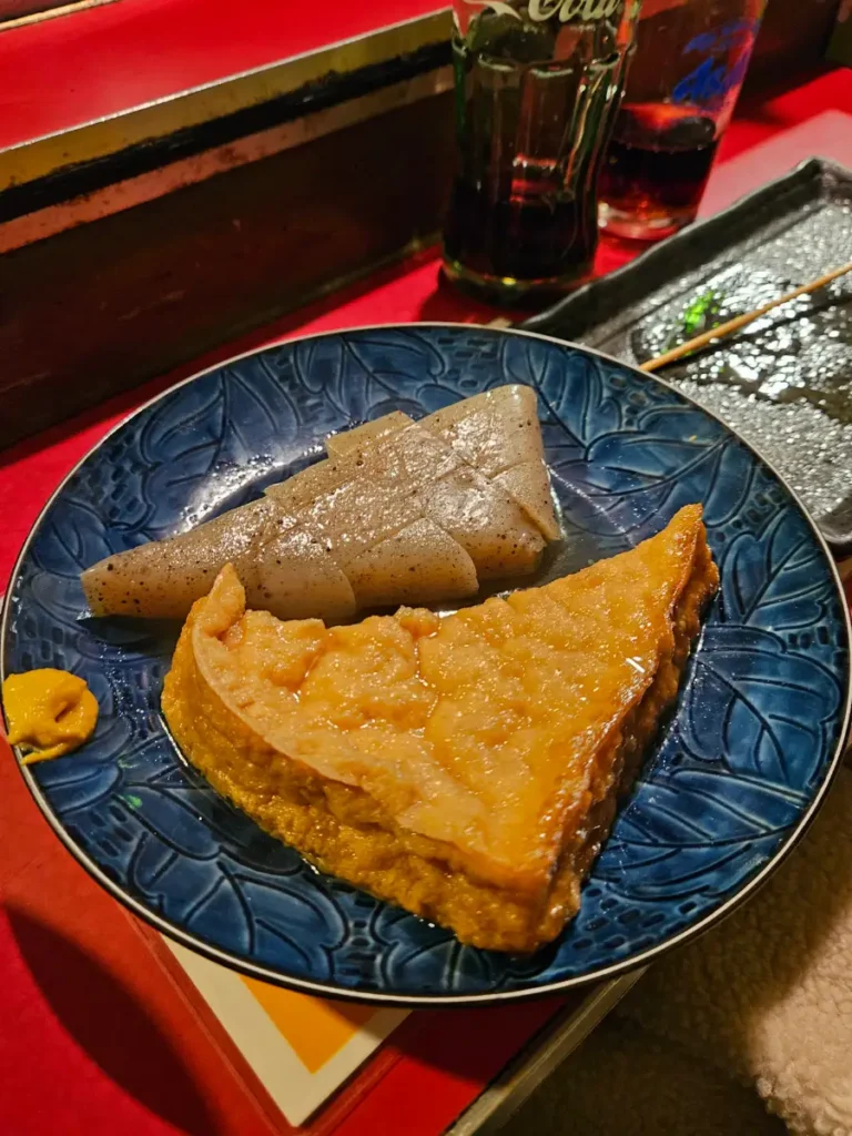 Glistening konjac and tofu oden on a blue plate with a dollop of spicy mustard on the side.