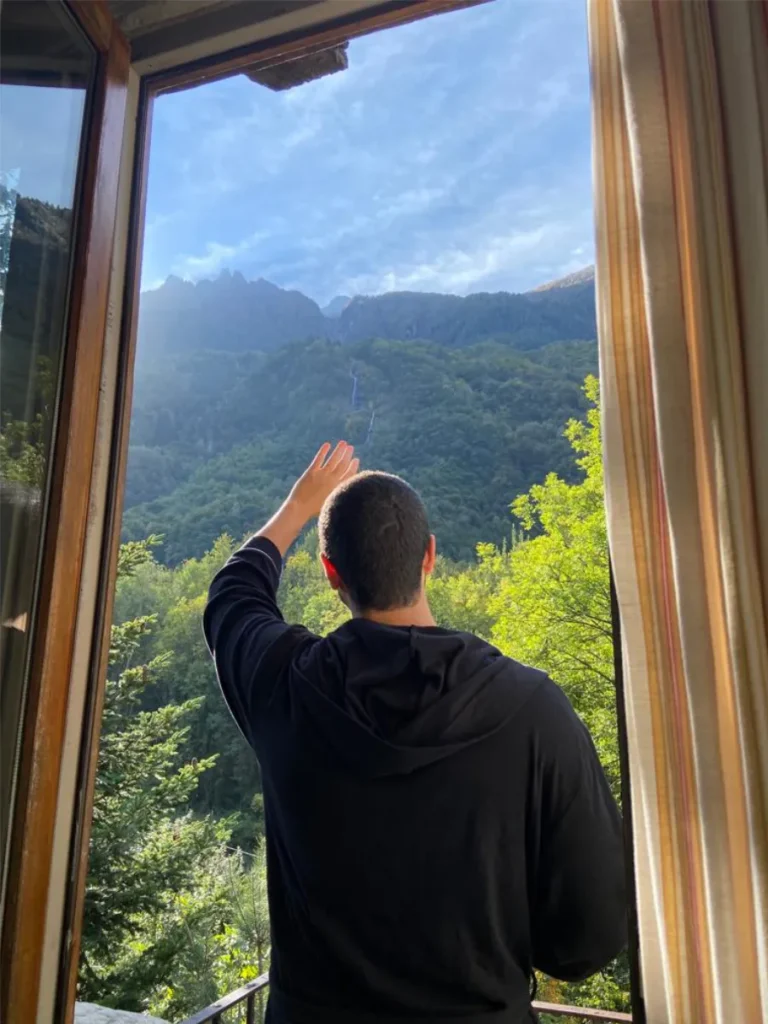 Tim drinking coffee looking up at the amazing view from a farmstay balcony of a waterfall and Italian mountain range.
