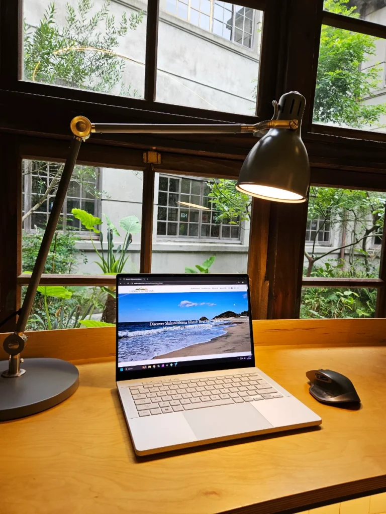 Aesthetic view of laptop, mouse, and lamp on a wooden desk looking out a window into a garden.