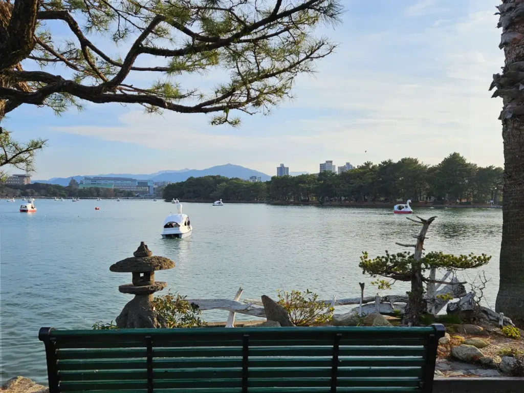 A scenic view of the lake at Ohori Park in Fukuoka, Japan, with white swan boats on the water, a stone lantern in the foreground, and a park bench overlooking the mountains in the distance.