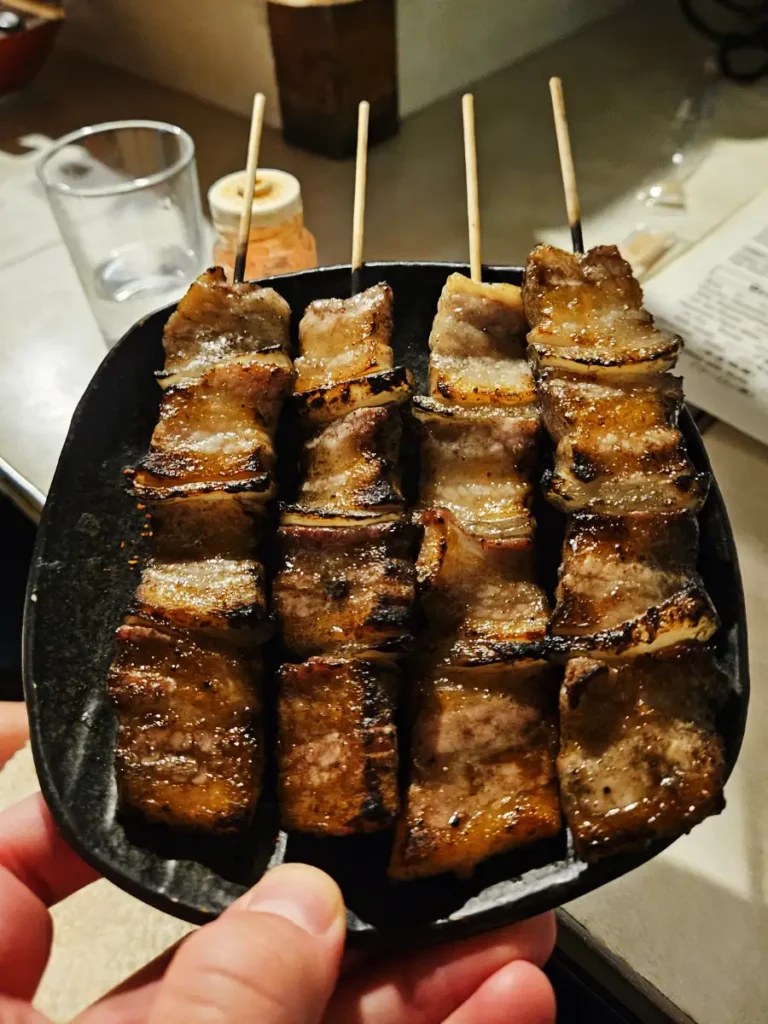 Four shining, crispy, pork belly and onion yakitori skewers on a black plate.