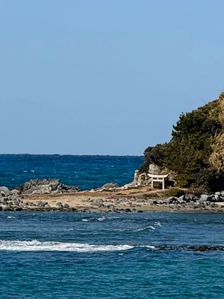 A white torii gate stands on a rocky shoreline on Shikanoshima Island, Fukuoka, with blue ocean waves in the foreground.