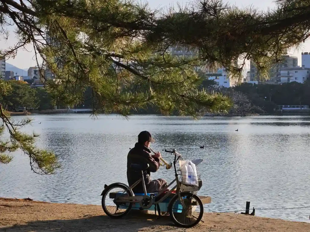 A man sitting next to his bicycle playing a trumpet overlooking Ohori Pond at Ohori Park.