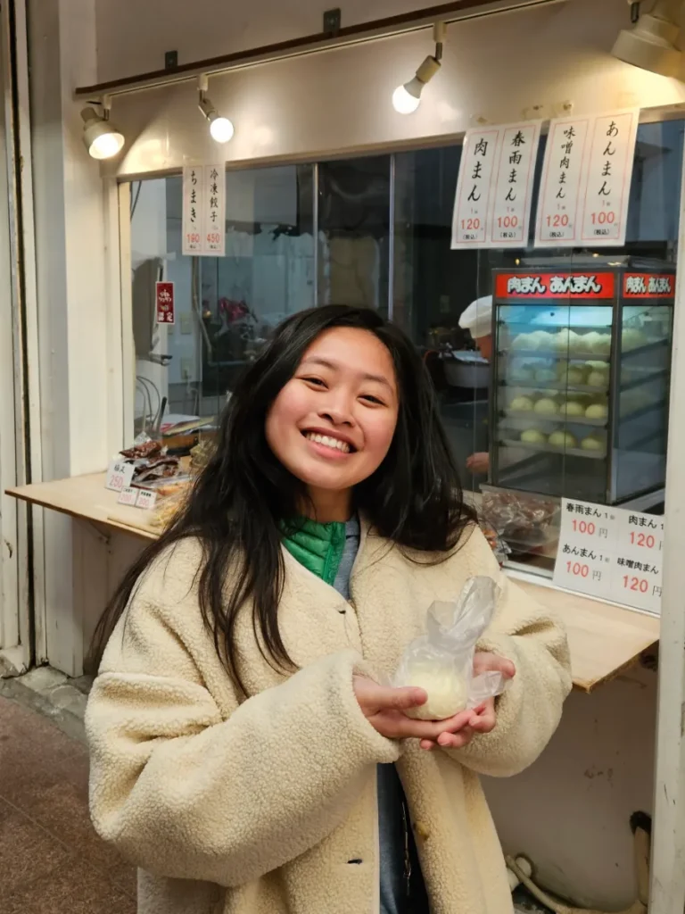 Zoe smiling holding a steamed bun in front of the shop that sold it.