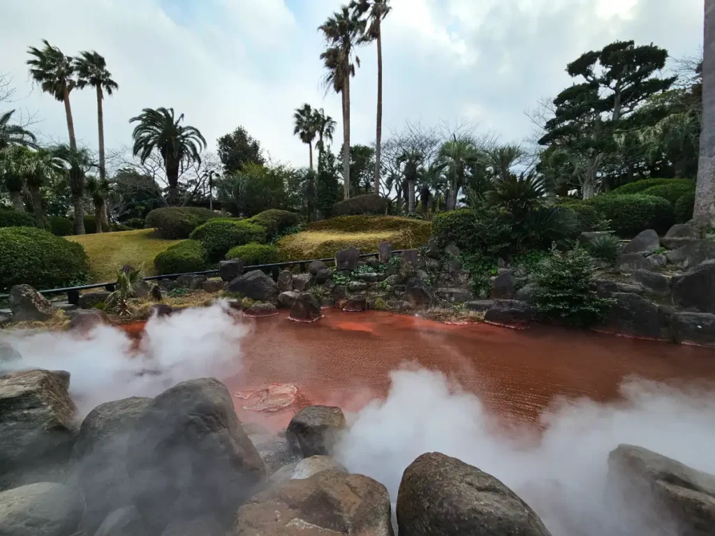 A view of Akaike, the secondary red thermal pond at Umi Jigoku. The water is a deep, rusty orange-red, with light steam rising from the surface and green foliage in the background.