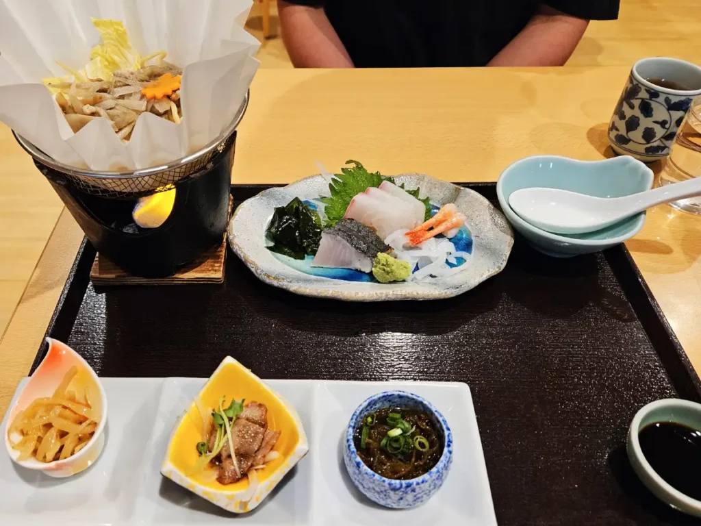 A Japanese set meal at Toyotsune restaurant, featuring raw fish, a pot of soup and vegetables, and assorted small side dishes.