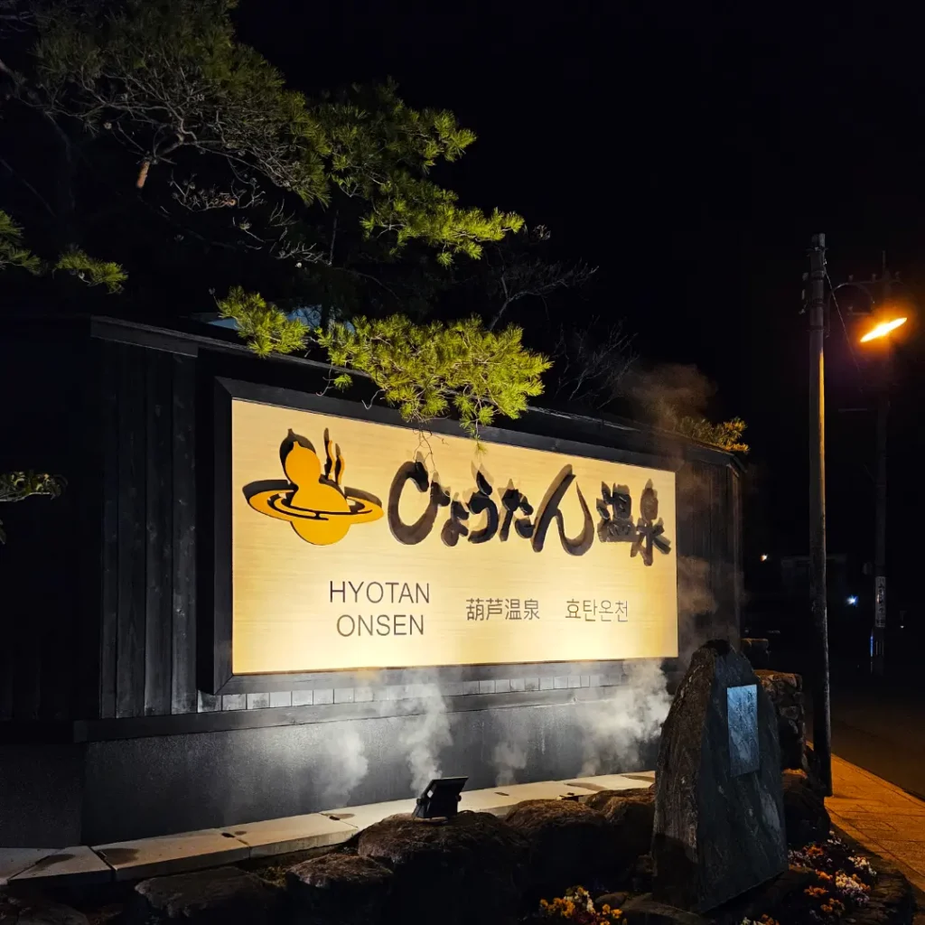 The entrance sign for Hyotan Onsen at night, illuminated by warm glowing lights. White steam rises from vents in the ground.