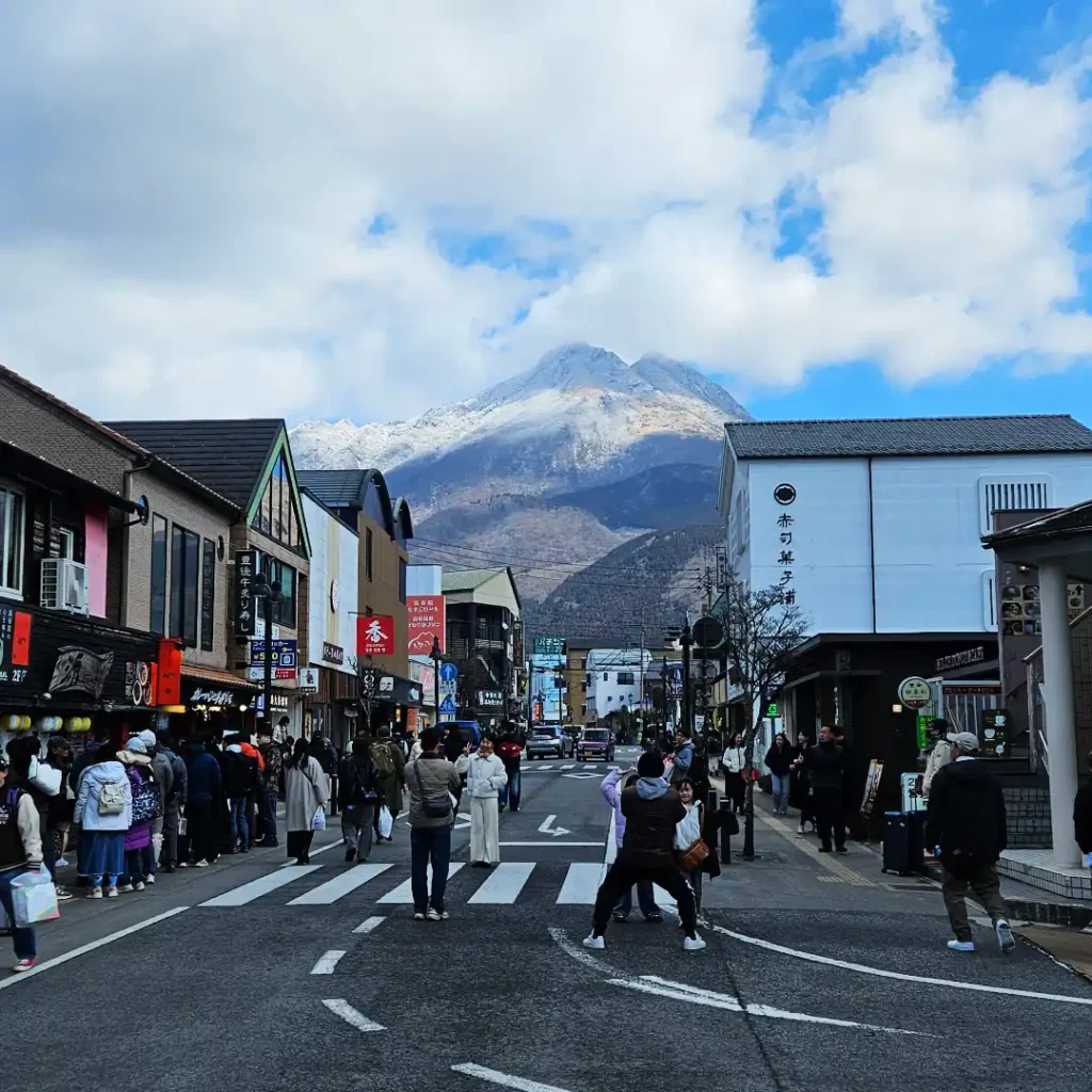 A busy street in Yufuin, with visitors posing for pictures and a snow-dusted Mount Yufu in the background.