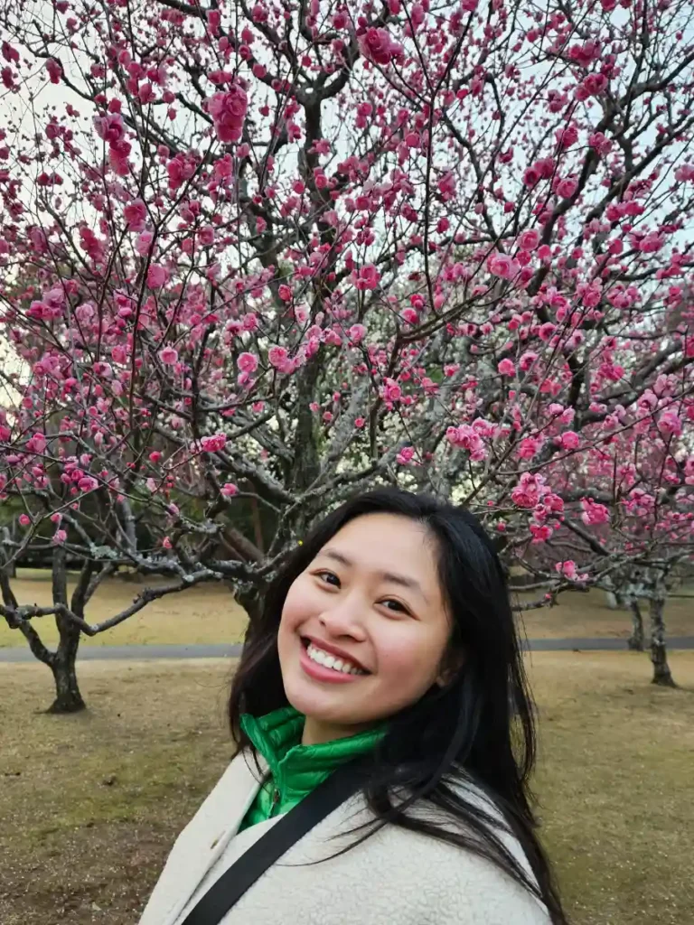 Zoe smiling in front of vibrant pink plum blossoms in Beppu Park.