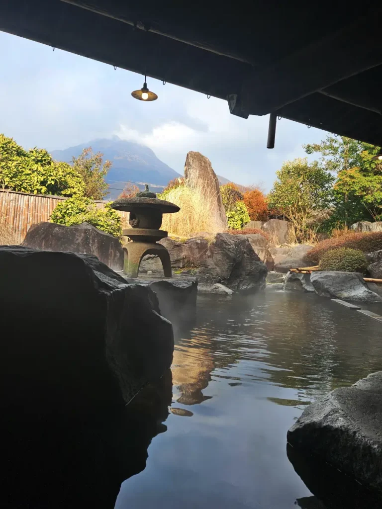 A serene outdoor hot spring bath at Ryoso Makibanoie in Yufuin. The steaming pool features large river stones, foliage, and a view of Mount Yufu.