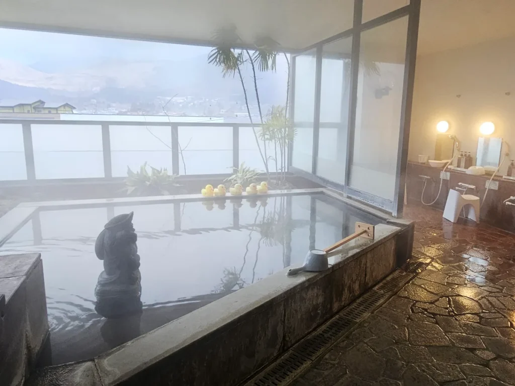 A misty semi-open-air hot spring bath with yellow rubber ducks on the edge of a stone tub.