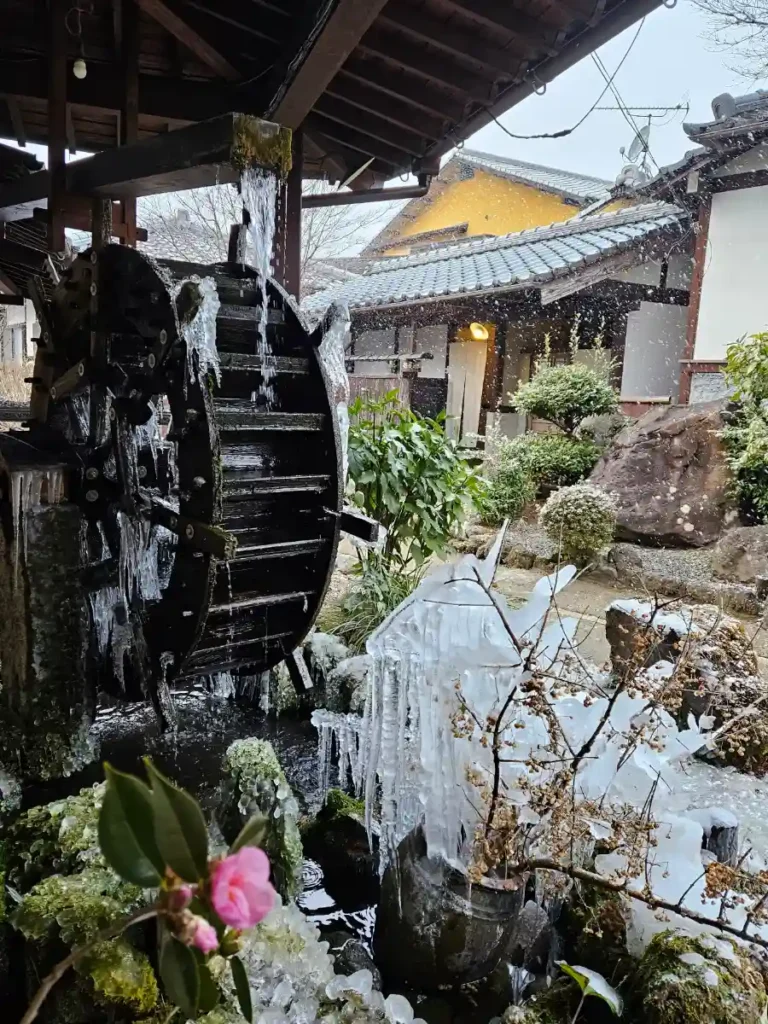A wooden water wheel in the garden at Ryoso Makibanoie. Fresh white snow is falling, while long icicles cling to the nearby garden plants and parts of the wheel.