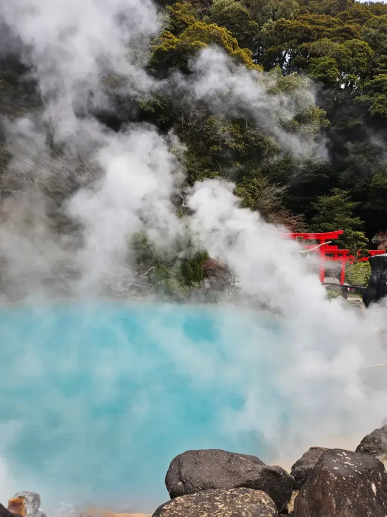 Bright red torii gates partially obscured by thick, swirling white steam rising from the boiling blue waters of Umi Jigoku in Beppu.