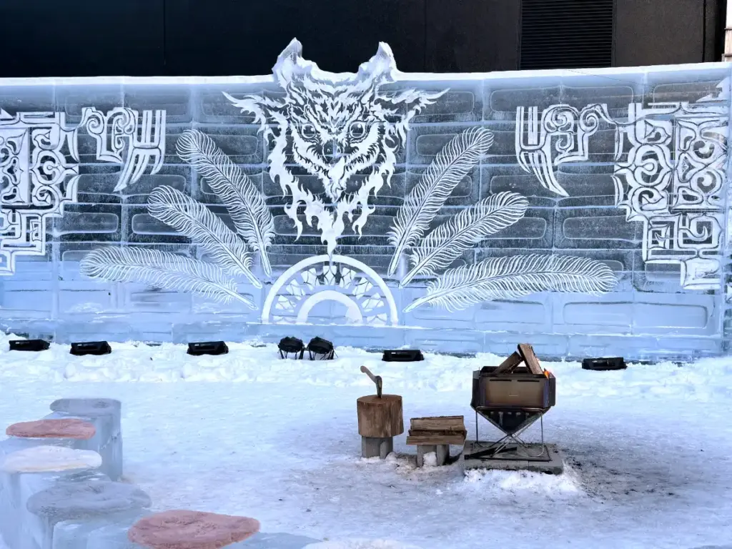 An ice wall carved with an owl and traditional indigenous Ainu motifs next to a fire pit with ice seating in Sapporo.