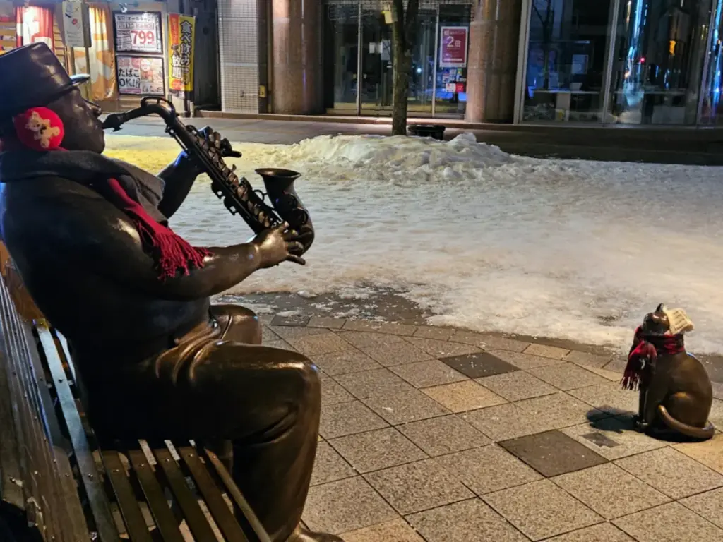 A bronze statue of a man playing a saxophone and a small cat, both dressed in scarves and earmuffs for a winter in Japan.