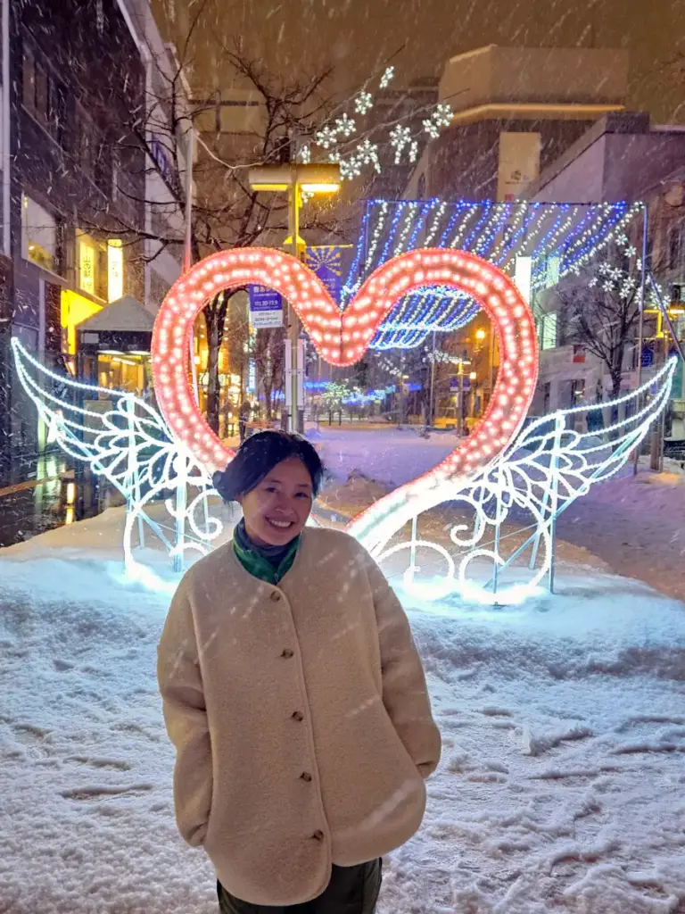 Zoe smiling in front of a glowing heart with angel wings and winter lights at the Heiwa Dori Shopping Park in Asahikawa.