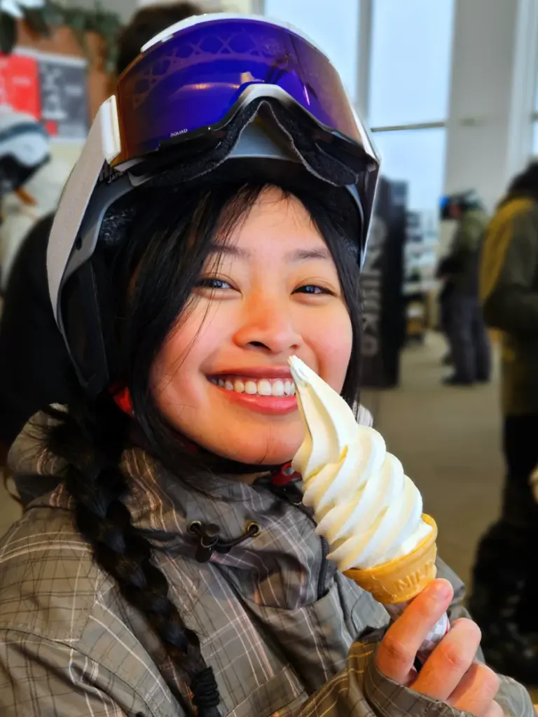 Zoe wearing a snowboarding helmet and goggles while smiling and eating a soft serve ice cream made with Hokkaido dairy.
