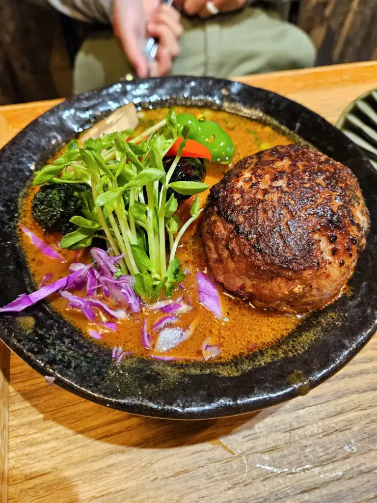 A steaming bowl of Hokkaido soup curry topped with a thick hamburger steak, broccoli, and greens to warm up during a freezing Japan winter.