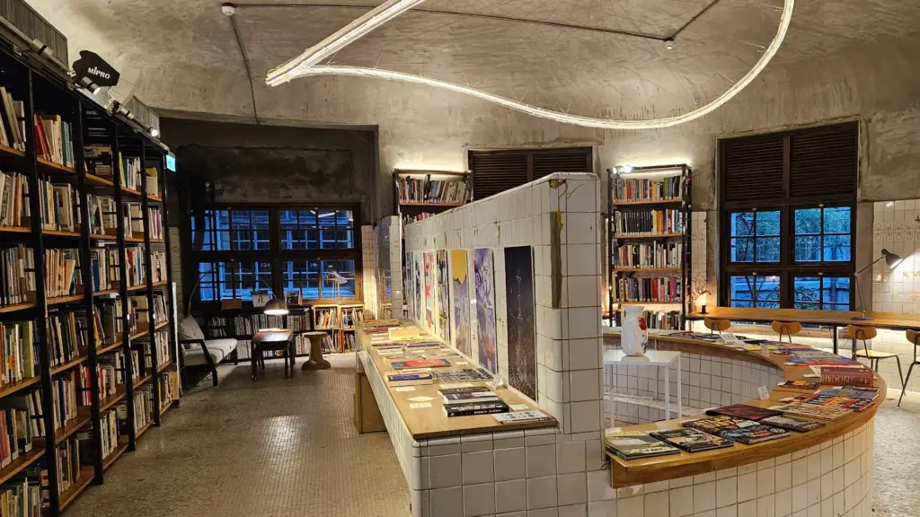 The interior of Not Just Library at Songshan Cultural and Creative Park in Taipei, Taiwan. The room features a curved white-tiled counter that was formerly a bathhouse tub, now filled with books. Dark wooden bookshelves line the walls under a high concrete ceiling.
