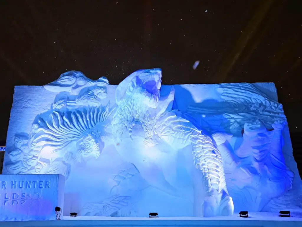 A massive, intricately carved snow sculpture of a roaring monster illuminated in bright blue light at night at the Sapporo Snow Festival.