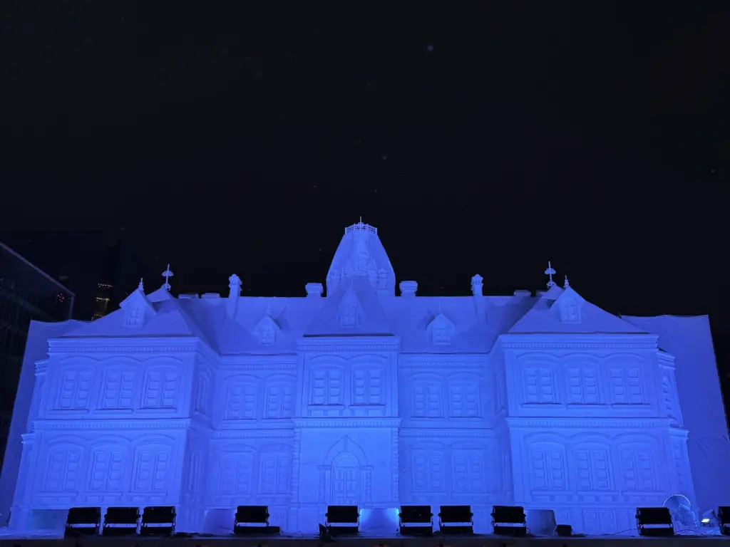 A massive, intricately carved snow sculpture of an elegant palace illuminated in bright blue light against the night sky at the Sapporo Snow Festival.