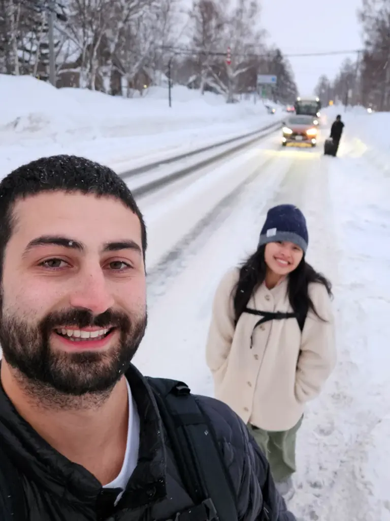 Tim and Zoe smiling while walking down a snowy road with their backpacks to reach their Niseko accommodation during a freezing Japan winter.