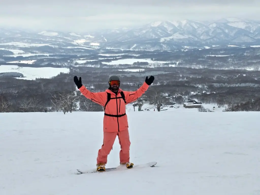 Tim standing on his snowboard with his arms outstretched, with a view of the mountain peaks behind him.