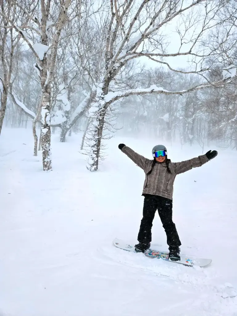 Zoe standing on her snowboard with her arms raised in the air, surrounded by snow-covered trees in the famous Niseko powder.