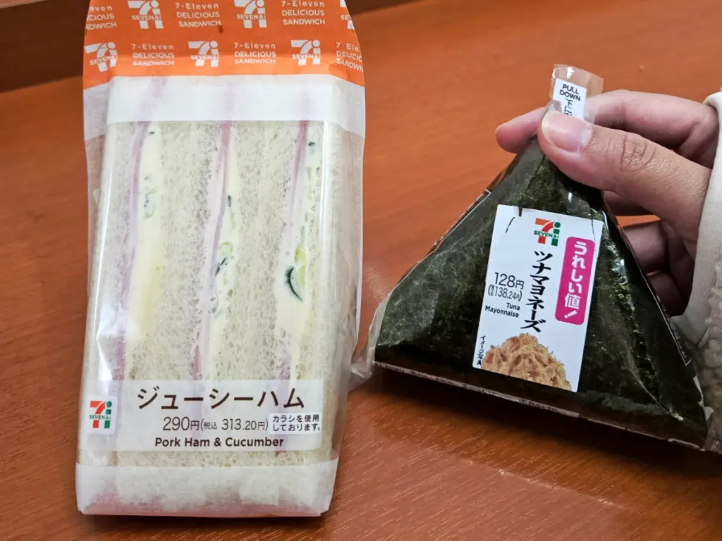 A close-up of convenience store snacks in Japan, showing a sandwich next to a wrapped tuna onigiri from 7-Eleven.