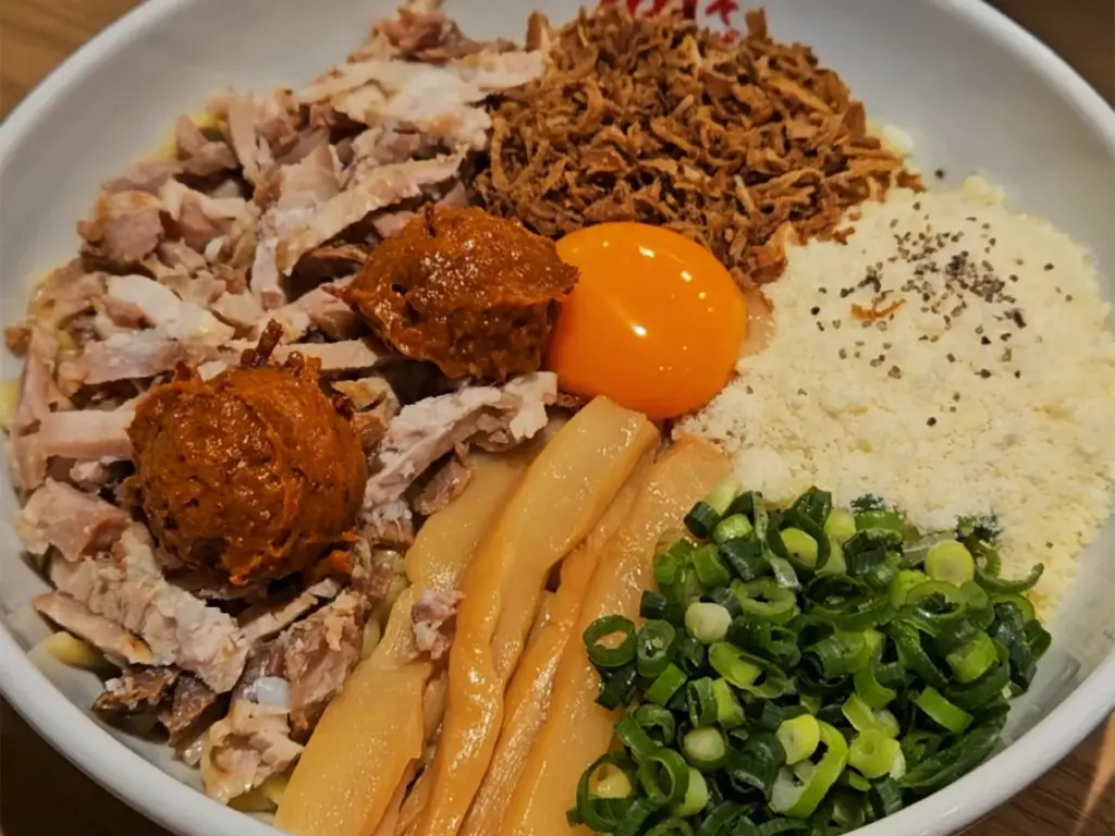 A colorful, packed bowl of abura soba noodles topped with chashu pork, green onions, bamboo shoots, fried onions, spices, and a bright yellow raw egg yolk.
