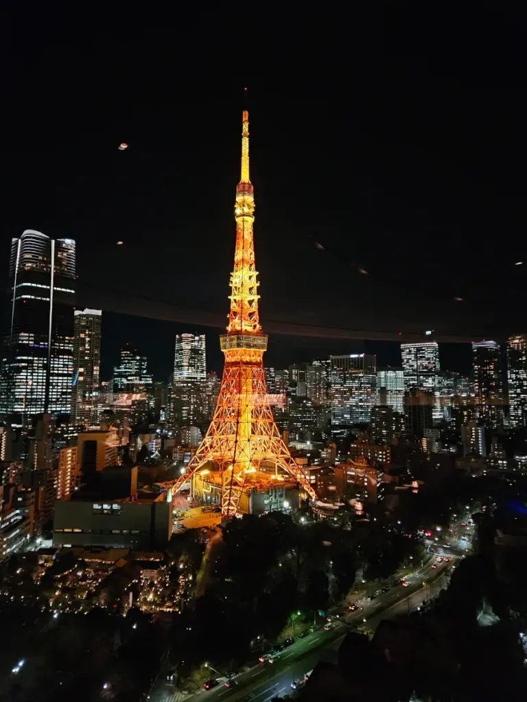 A brilliant night view of the illuminated Tokyo Tower surrounded by glowing city skyscrapers, taken through the glass window of the Sky Lounge in Tokyo.