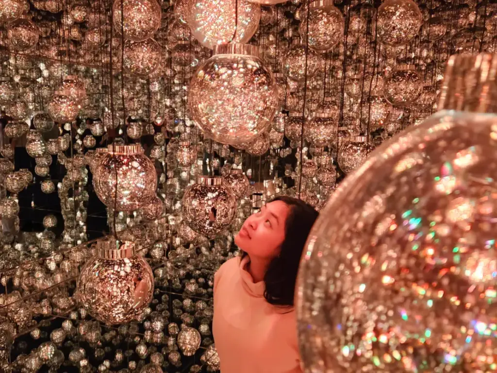 Zoe inside a mirrored room filled with hundreds of hanging glass spheres glowing with warm, sparkling copper light in the Bubble Universe at teamLab Borderless.