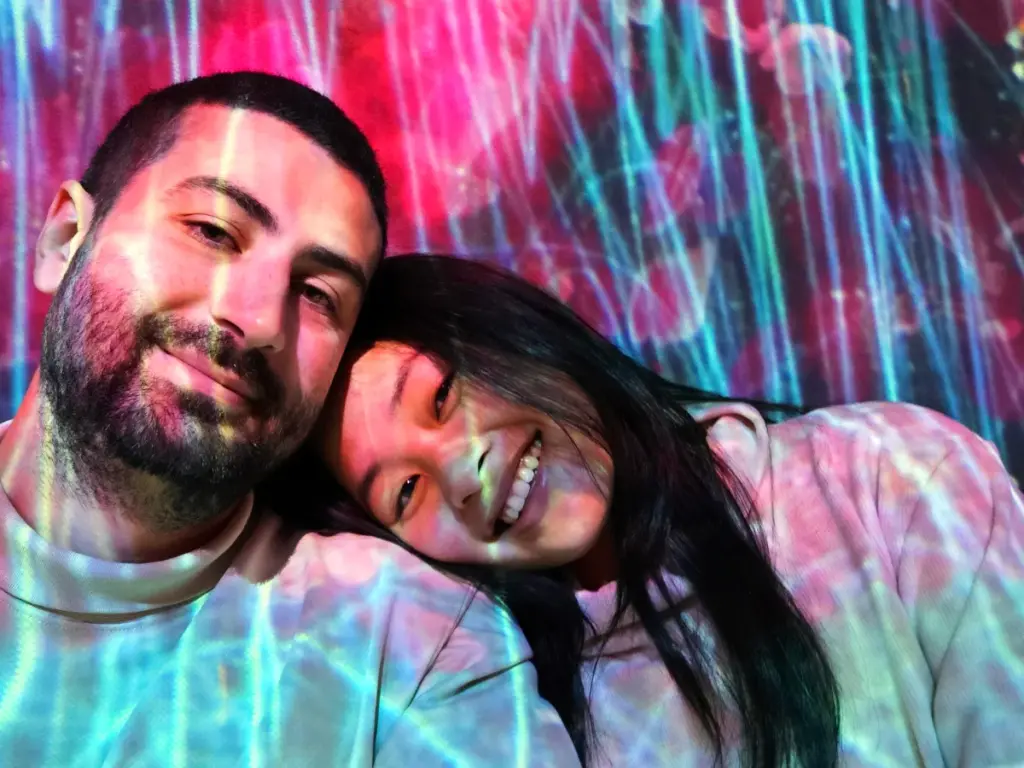 Zoe and Tim smiling together, illuminated by vibrant, neon light projections across their faces inside teamLab Borderless in Tokyo.