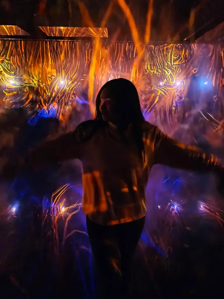 Zoe standing with her arms outstretched in a dark room, surrounded by vibrant digital projections of flames on smoke at teamLab Borderless Tokyo.