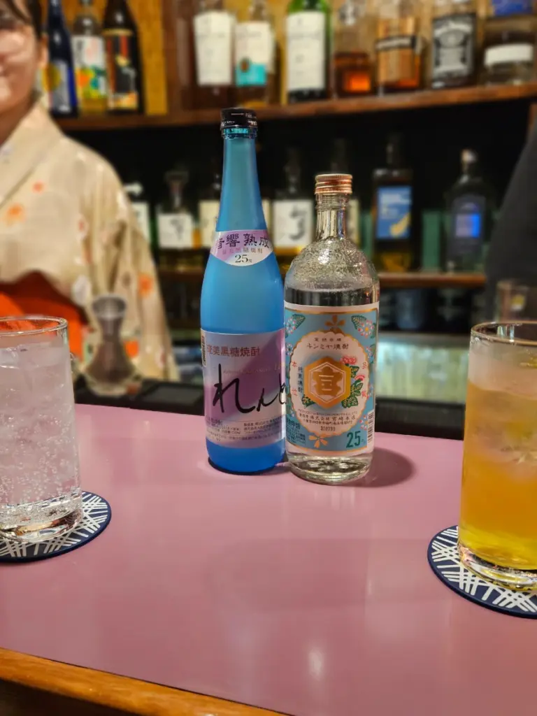 Two bottles of Japanese liquor and two cocktail glasses resting on a pink bar counter at a tiny establishment in Tokyo's Golden Gai, with a bartender wearing a kimono in the background.