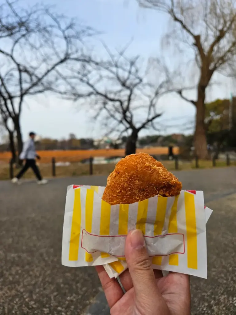 Holding Famichiki fried chicken in its signature yellow-and-white striped paper bag, standing on a path in Ueno Park, Tokyo. The background shows bare winter trees and Shinobazu Pond in the distance.