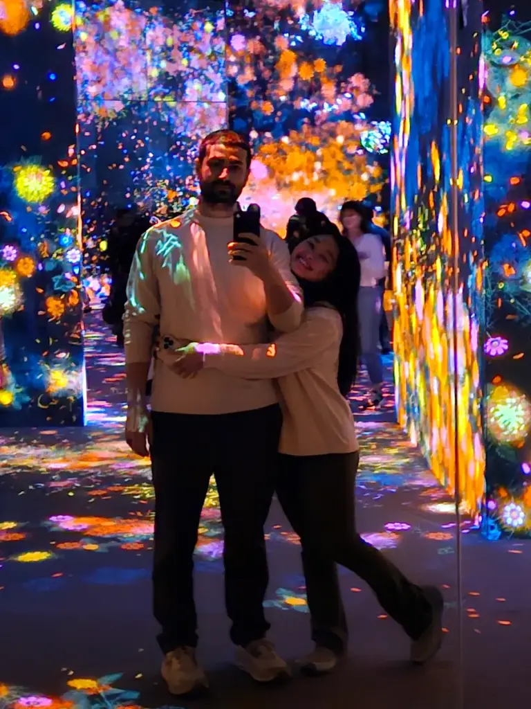 A couple hugging in a dark, mirrored room surrounded by brightly colored, interactive digital flower projections at teamLab Borderless.