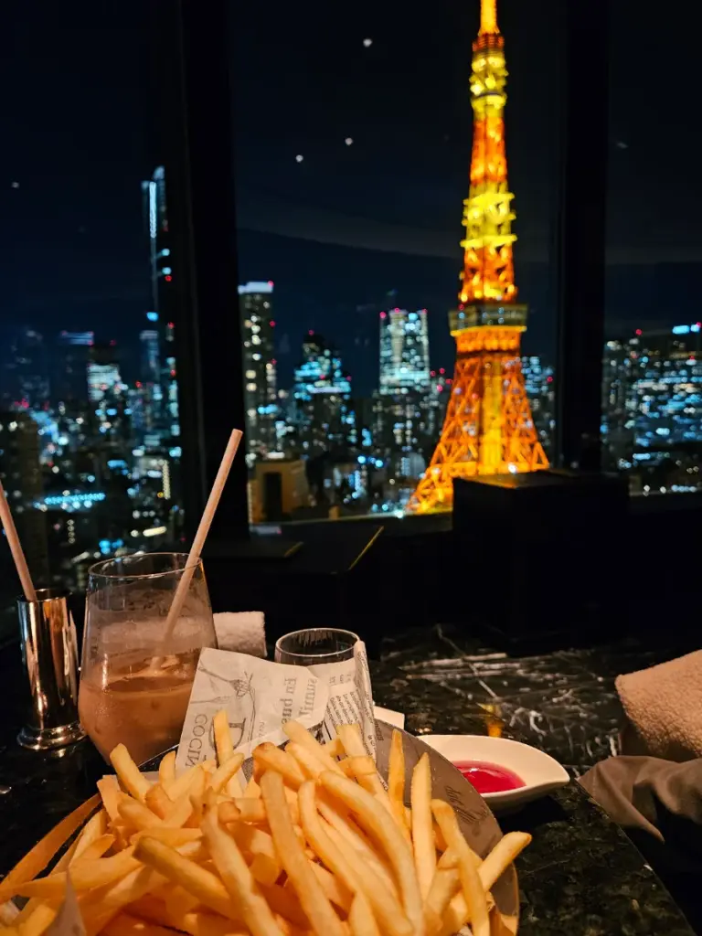 A basket of french fries and cocktails on a table at the Sky Lounge Prince Park Tower Tokyo, with a stunning, brightly illuminated night view of the Tokyo Tower shining right outside the window.