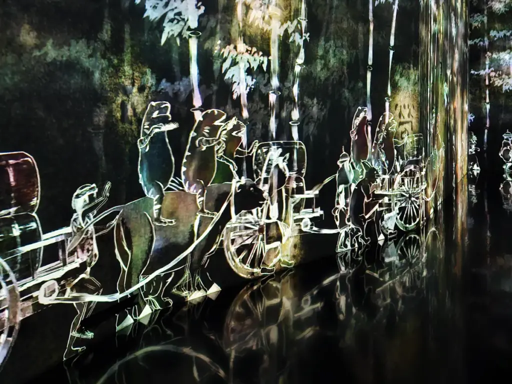 A digital art projection on a dark wall showing glowing, animated frogs walking in a procession alongside a cart at teamLab Borderless Tokyo.