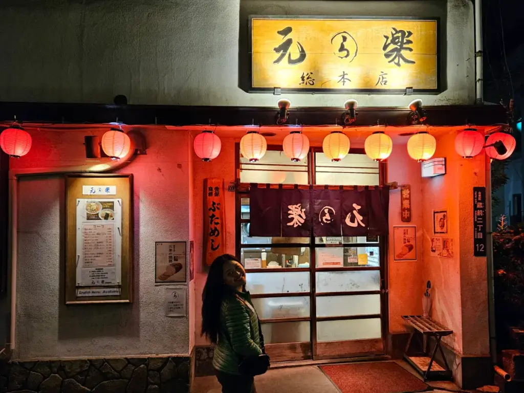 Zoe standing happily in front of the rustic exterior of the Genraku ramen shop in Asakusa at night, illuminated by a row of glowing red paper lanterns.