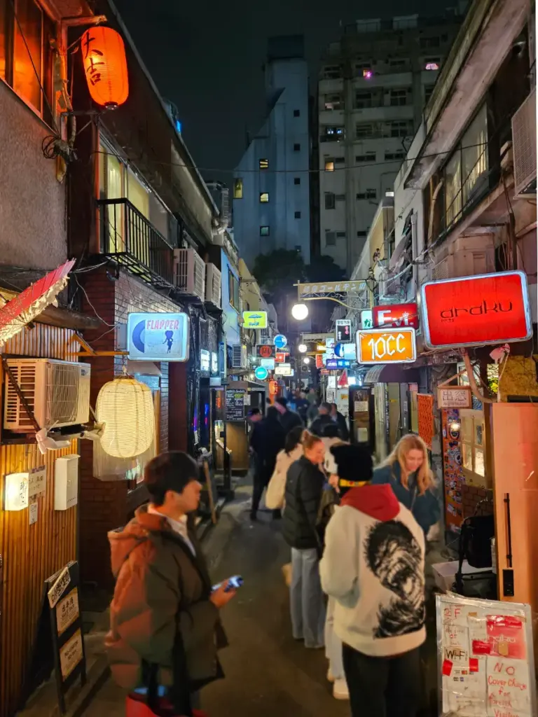 A busy, narrow alleyway in Tokyo's Golden Gai district at night, filled with people walking past brightly lit, colorful neon bar signs.