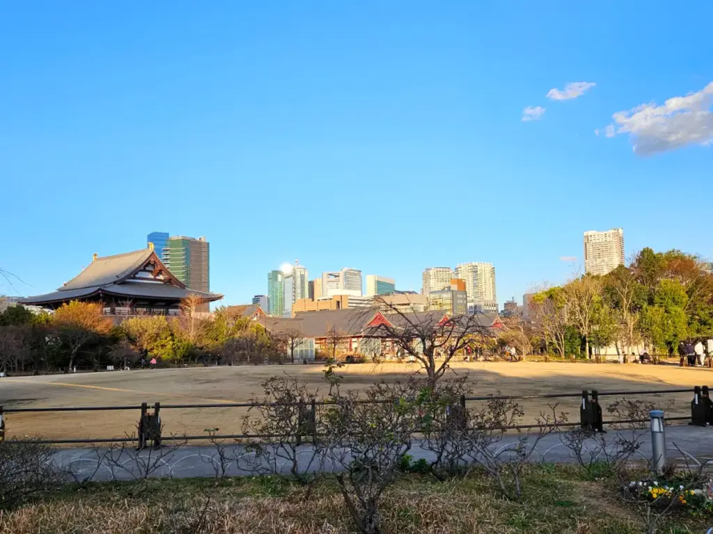 A view of a wide field at Shiba Park in Tokyo with traditional temple rooflines on the left and modern city skyscrapers in the background under a clear blue sky.