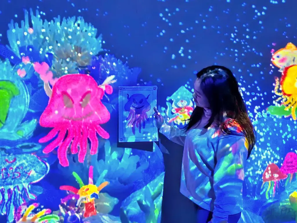 Zoe holding up her colored drawing of a pink jellyfish next to a dark blue wall, where her exact drawing is animated and projected swimming among other colorful digital sea creatures in the Sketch Ocean exhibit.