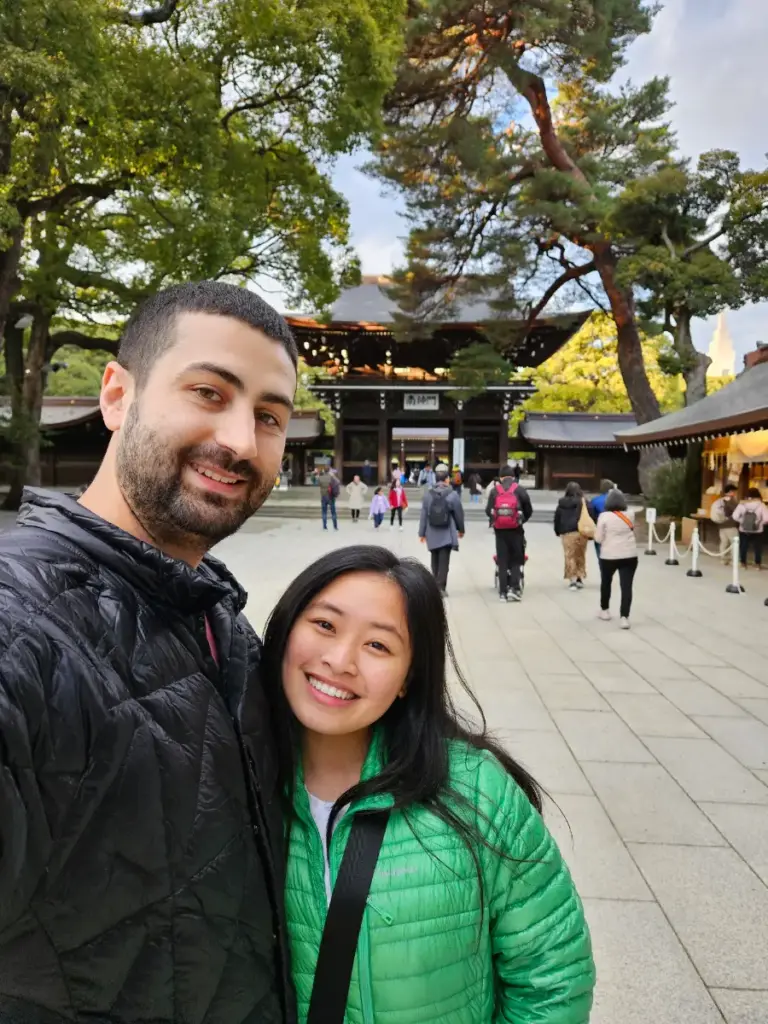 A selfie of Zoeand Tim at Meiji Jingu shrine, smiling in a paved courtyard with traditional Japanese wooden shrine buildings and a torii gate visible behind them.