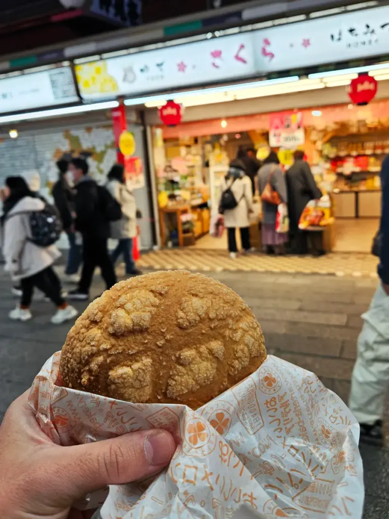 Holding a fresh Japanese melonpan sweet bread in Asakusa, Tokyo. The melonpan has a crispy, sugary crust and is wrapped in decorative paper. In the background, there is a slightly blurred traditional street market at dusk.